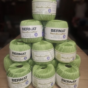 May include: A collection of light green Bernat Handicrafter crochet thread balls stacked on a wooden surface. Each ball has a white label with black text. The labels read "Bernat Handicrafter Crochet Thread".