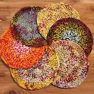 May include: A collection of seven colorful, round crochet trivets. Each trivet features a spiral design with a mix of warm colors, including red, orange, yellow, and green. The trivets are arranged on a wooden surface.