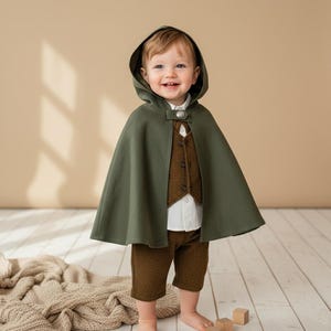 Puede incluir: Un beb&eacute; con una capa con capucha verde, chaleco marr&oacute;n, camisa blanca y pantalones cortos marrones. La capa se abrocha con un bot&oacute;n decorativo. El atuendo est&aacute; dise&ntilde;ado para un ni&ntilde;o y es adecuado para un disfraz u ocasi&oacute;n especial.