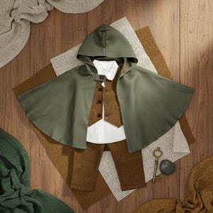 Puede incluir: Un conjunto infantil con capa con capucha verde oliva, chaleco marr&oacute;n, camisa blanca y pantal&oacute;n marr&oacute;n. Una peque&ntilde;a llave antigua y una piedra est&aacute;n colocadas cerca.