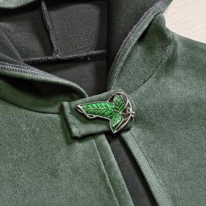 Puede incluir: Primer plano de una capa con capucha verde oscuro con un cierre decorativo en forma de hoja. El cierre es plateado con detalles de esmalte verde. La capa tiene una textura suave similar a la gamuza.