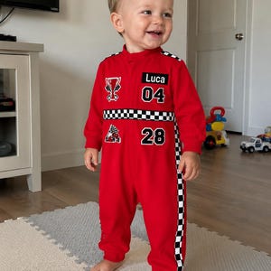 May include: A red long-sleeve jumpsuit featuring a black and white checkered stripe across the chest and down the sides. The garment displays the name Luca, the numbers 04 and 28, a trophy graphic, and a checkered flag graphic in white and black.