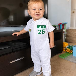 May include: A white jumpsuit featuring a green and white logo, the name "JETT", and the number "25" on the chest. The outfit is paired with grey trainers. The jumpsuit is designed for a toddler.