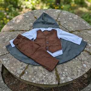 Puede incluir: Un conjunto de ropa de beb&eacute; que incluye un chaleco marr&oacute;n con tres botones, pantalones marrones, una camisa blanca con cuello y una capa con capucha gris. El conjunto est&aacute; dispuesto sobre una superficie de piedra. La ropa es de tela texturizada.