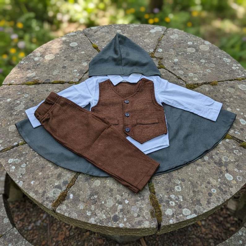 Puede incluir: Un conjunto de ropa de beb&eacute; que incluye un chaleco marr&oacute;n con tres botones, pantalones marrones, una camisa blanca con cuello y una capa con capucha gris. El conjunto est&aacute; dispuesto sobre una superficie de piedra. La ropa es de tela texturizada.