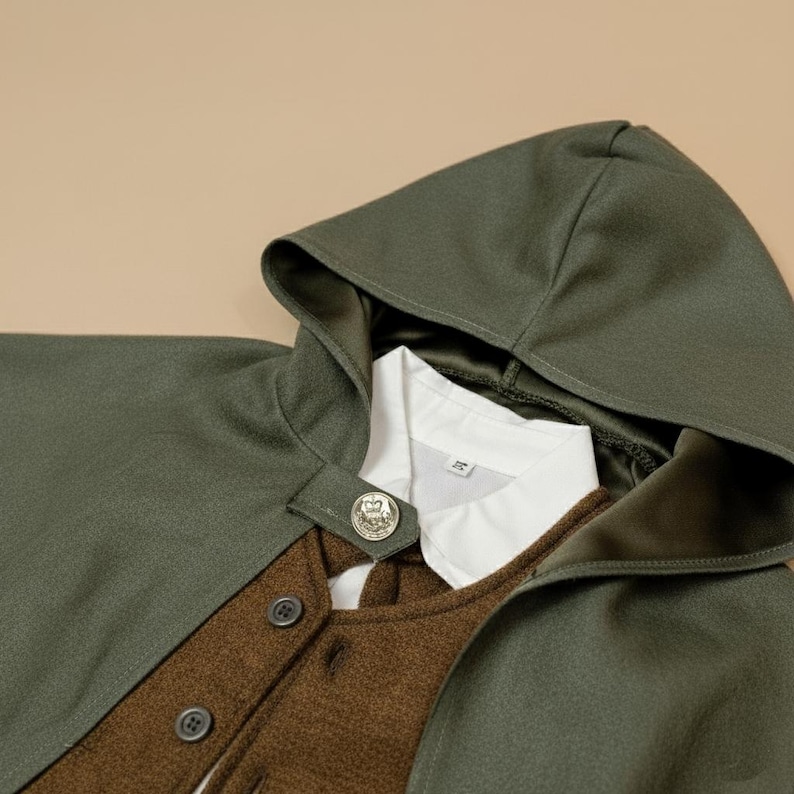 Puede incluir: Una capa con capucha verde oliva con un chaleco marr&oacute;n y una camisa blanca con cuello. La capa tiene un cierre de bot&oacute;n y un emblema decorativo. El atuendo es en capas y parece estar hecho de lana o un tejido similar.