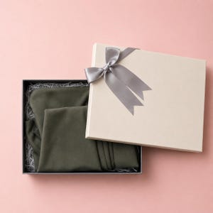 Puede incluir: Una caja de regalo abierta con tapa color crema atada con una cinta de raso gris. Dentro de la caja negra hay una prenda verde oliva doblada, sobre papel triturado, sobre un fondo rosa.