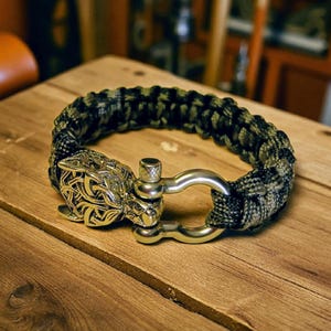 Könnte beinhalten: Ein geflochtenes Paracord-Armband in Schwarz-, Grün- und Beigetönen. Das Armband hat einen dekorativen goldfarbenen Metallverschluss mit einem detaillierten Wolfskopf-Design und einen silberfarbenen Schäkelverschluss. Das Armband wird auf einer Holzoberfläche präsentiert.