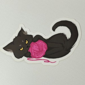 以下が含まれることがあります： 黄色い目をした黒い猫が横向きに寝て、ピンクの毛糸玉を持っています。