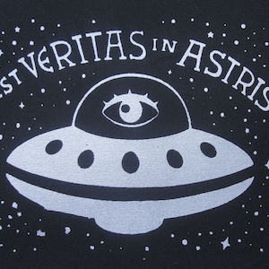 Può includere: Un'illustrazione in bianco e nero di un disco volante con un occhio al centro. Il testo "EST VERITAS IN ASTRIS" è scritto intorno al disco.