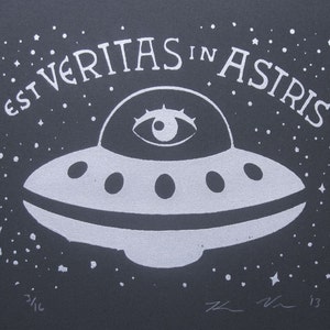 May include: A black and white illustration of a flying saucer with an eye in the center. The text "EST VERITAS IN ASTRIS" is written around the saucer.