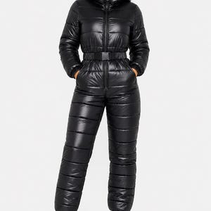 Puede incluir: Un mono acolchado de color negro con mangas largas, cuello alto, capucha incorporada y cierre de cremallera frontal. La prenda incluye un cinturón negro a la cintura con hebilla de plástico y puños elásticos en las muñecas y los tobillos.