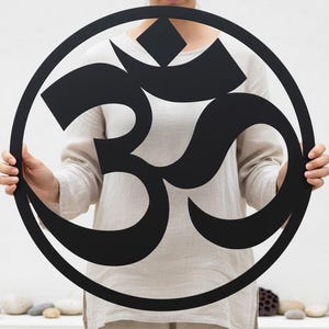 Könnte beinhalten: Ein großes, schwarzes Om-Symbol aus Metall, das von einer Person gehalten wird. Das Symbol ist kreisförmig mit einem stilisierten Design. Der Hintergrund ist weiß mit ein paar Steinen unten.