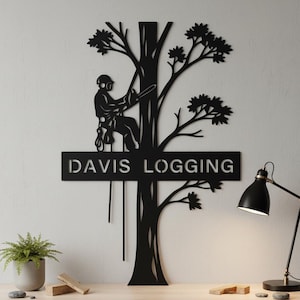Op de afbeelding: Een zwart metalen wanddecoratie met het silhouet van een boomklimmer die een kettingzaag gebruikt, geïntegreerd met een horizontaal rechthoekig bord met de tekst DAVIS LOGGING in een uitgesneden schreefloos lettertype.