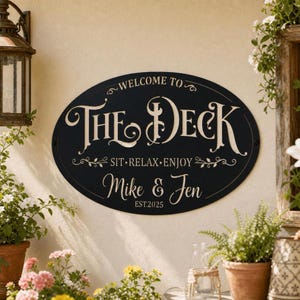 Könnte beinhalten: Ein ovales schwarzes Metallschild mit weißer Schrift, die WELCOME TO THE DECK, SIT RELAX ENJOY, Mike & Jen und EST.2025 lautet. Das Schild ist mit dekorativen Schnörkeln versehen und an einer Wand mit einer Laterne und Pflanzen montiert.