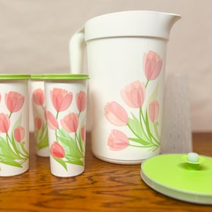 May include: A white pitcher and four matching tumblers with a pink tulip design. The tumblers have green lids. A green lid and a clear plastic cup are also included. The set is designed for serving beverages.