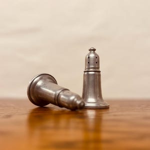 May include: Two antique silver salt and pepper shakers. One shaker is upright, the other is lying on its side. The shakers have a classic design with a flared base and a small, rounded top. The salt shaker has holes on the top.