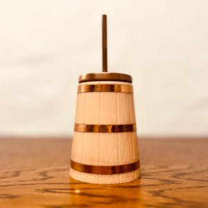 May include: A wooden butter churn with copper-colored bands and a matching handle. The churn has a light wood finish and a classic barrel shape. The copper bands add a decorative touch to the kitchenware.