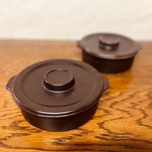 May include: Two dark brown ceramic lidded pots. Each pot has a round shape with a lid featuring a central knob. The pots are on a wooden surface, with one pot in the foreground and the other slightly behind it.