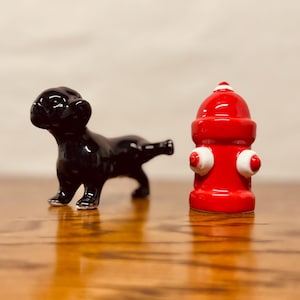 May include: A black ceramic dog figurine and a red ceramic fire hydrant with white accents. The dog is in a playful pose, and the hydrant has a glossy finish. The figurines are on a wooden surface.