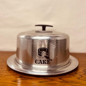 May include: A vintage silver cake carrier with a domed lid and a round base. The lid has a black handle and a graphic of a cake with the word "CAKE" below it. The carrier is made of metal and sits on a wooden surface.