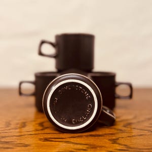 May include: A stack of dark brown ceramic mugs with square handles. One mug is flipped over, revealing the white rim and the text "M. DOVESTONE ENGLAND" in a circular pattern. The mugs are on a wooden surface.
