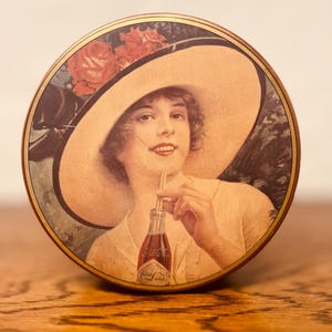 May include: Round vintage Coca-Cola tin featuring a woman in a large hat, sipping from a Coca-Cola bottle. The tin has a gold rim and a floral design. The woman is wearing a light-colored dress.
