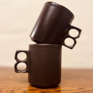 May include: Two dark brown ceramic mugs stacked on a wooden surface. Each mug has a unique handle design with two finger holes. The mugs are cylindrical with a smooth, matte finish, and are set against a neutral background.