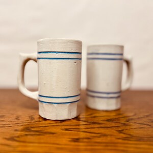 May include: Two off-white ceramic mugs with blue horizontal stripes. Each mug has a handle and a cylindrical shape. The mugs are displayed on a wooden surface.