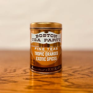 May include: A gold-colored tin of Boston Tea Party fine teas, flavored with tropic oranges and exotic spices. The label features the text "Boston Tea Party" in a white banner. The tin is sitting on a wooden surface.