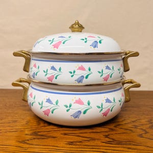 May include: Two white enamel pots with lids, stacked on top of each other. Each pot features a blue stripe, gold handles, and a floral pattern with pink and blue flowers. The lids have gold finials.