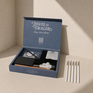 Könnte beinhalten: Ein blauer Set-Karton mit der Aufschrift Jewel Dealer clay into silver und einem QR-Code, der einen kleinen Gasbrenner, eine schwarze rechteckige Platte, ein Silberton-Päckchen und ein strukturiertes Werkzeug enthält. Fünf weiße Pinsel liegen daneben.