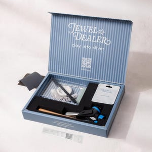 Könnte beinhalten: Ein blau-weiß gestreifter Set-Kasten mit der Aufschrift Jewel Dealer clay into silver, der einen Gasbrenner, ein Päckchen Silberton, einen Ringgrößenmesser, ein Schleifpad und Metallwerkzeuge enthält. Der Deckel zeigt einen QR-Code.