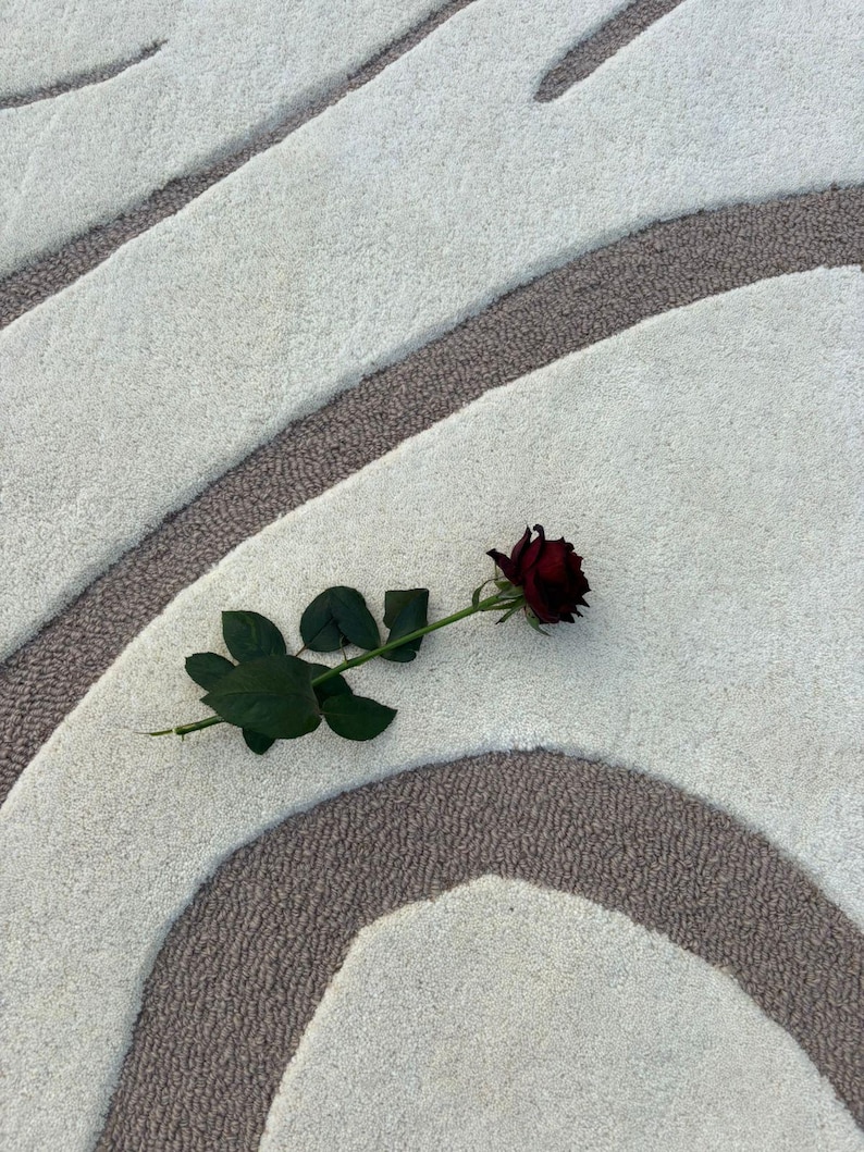 May include: A deep red rose with green foliage lies on a patterned rug. The rug's design features curved lines in white and brown, creating a soft, textured surface. The rose is in full bloom, with its petals fully open.