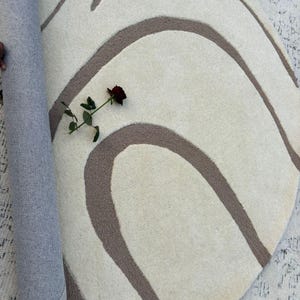 May include: A cream-coloured, round rug featuring a brown, wavy design. A rolled section of the rug shows a blue backing. A dark red rose rests on the rug's surface, adding a splash of colour.