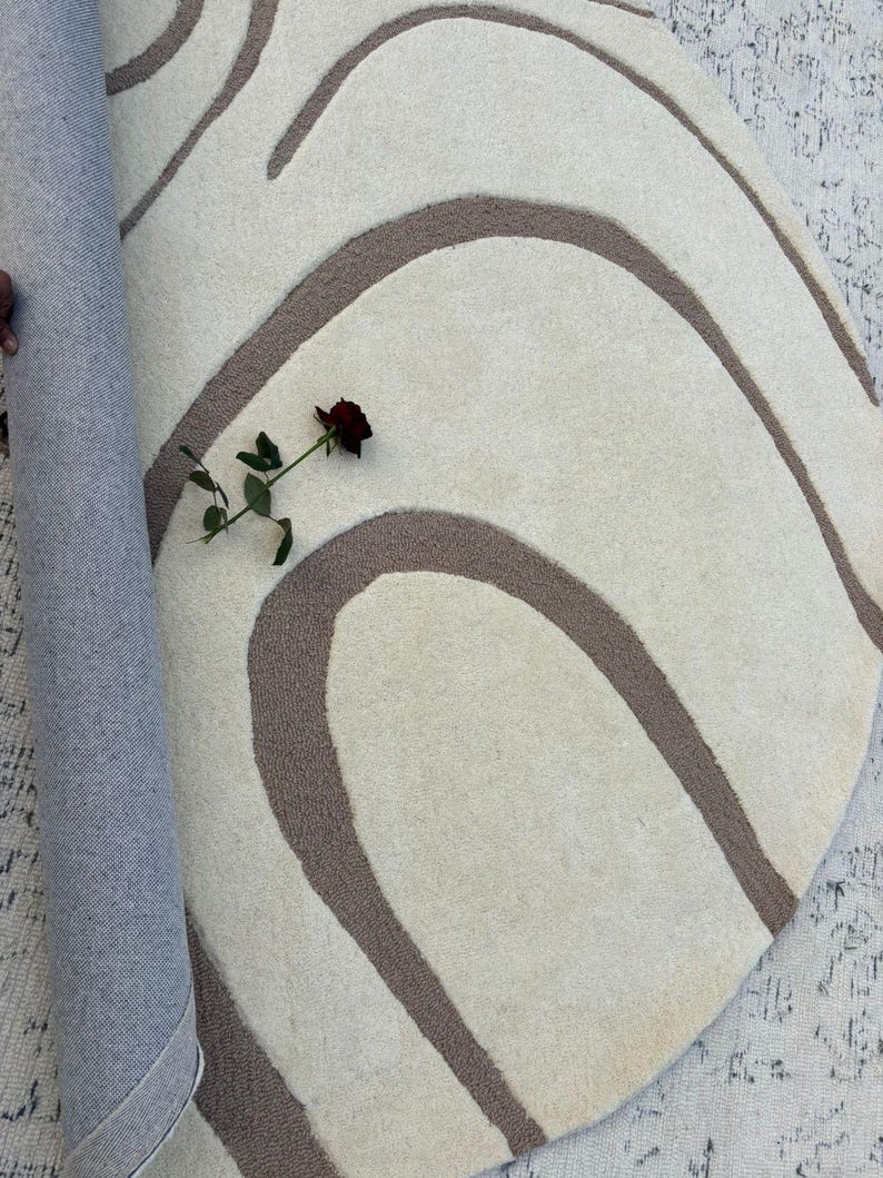 May include: A cream-coloured, round rug featuring a brown, wavy design. A rolled section of the rug shows a blue backing. A dark red rose rests on the rug's surface, adding a splash of colour.