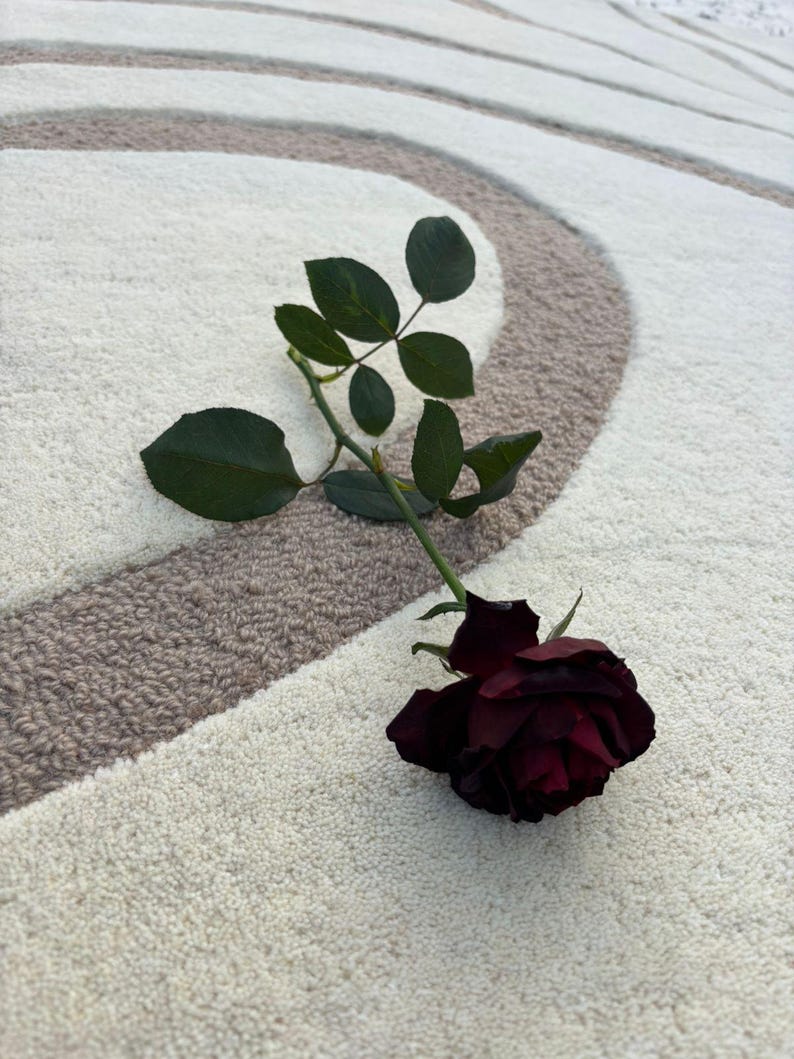 May include: A deep red rose with green leaves rests on a cream rug with a brown and cream circular design. The rose is fully open, displaying its rich colour and delicate petals.