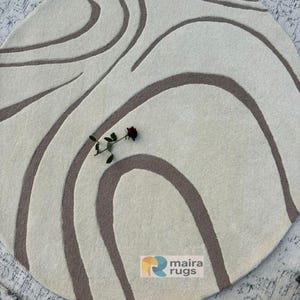 May include: A circular, cream-coloured rug featuring a brown, flowing, abstract pattern. A dark red rose rests on the rug. The rug has the text "maira rugs" on it.