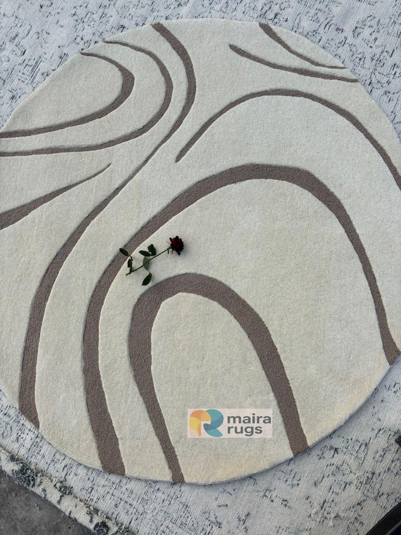 May include: A circular, cream-coloured rug featuring a brown, flowing, abstract pattern. A dark red rose rests on the rug. The rug has the text "maira rugs" on it.