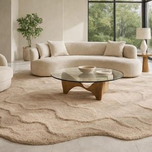 Op de afbeelding: Een moderne woonkameropstelling met een gebogen beige bank met bijpassende kussens, een ronde glazen salontafel en een beige tapijt met een organische vorm. Een beige fauteuil en een bijzettafel met een lamp maken de scène compleet.
