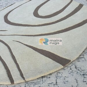 May include: Oval rug featuring a cream backdrop with dark brown, curved lines. The rug has a soft texture and a modern design. The logo "maira rugs" is visible on the rug.