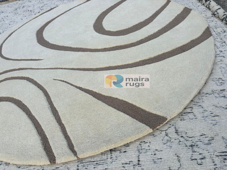 May include: Oval rug featuring a cream backdrop with dark brown, curved lines. The rug has a soft texture and a modern design. The logo "maira rugs" is visible on the rug.