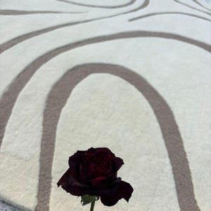May include: A circular, cream-coloured rug featuring a brown, curved design. A dark red rose with green leaves is in the foreground. The rug's pattern consists of overlapping arches and lines, giving it a textured look.