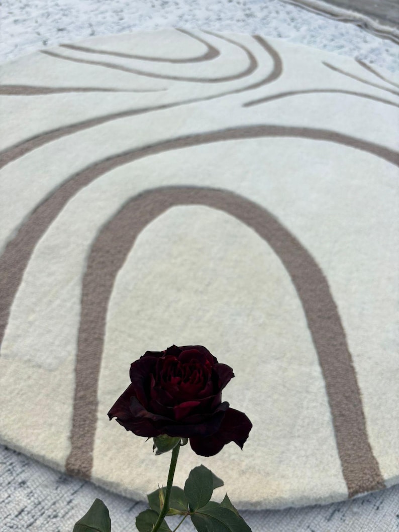 May include: A circular, cream-coloured rug featuring a brown, curved design. A dark red rose with green leaves is in the foreground. The rug's pattern consists of overlapping arches and lines, giving it a textured look.