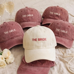 Op de afbeelding: Een verzameling baseballpetten in bordeauxrood en crème. De bordeauxrode petten zijn geborduurd met "THE GIRLS" in wit, terwijl de crèmekleurige pet "THE BRIDE" in rood heeft. Ideaal voor een vrijgezellenfeest.