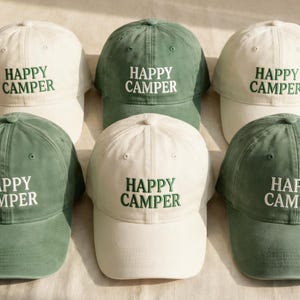Op de afbeelding: Een verzameling baseballpetten in wit en groen. Elke pet heeft de geborduurde tekst "HAPPY CAMPER" in een contrasterende kleur. De petten zijn netjes gerangschikt en tonen hun ontwerp en stijl.