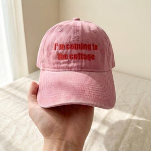 Peut inclure: Casquette de baseball rose poudré avec le texte brodé "I'm coming to the cottage" en rouge. La casquette a une visière incurvée et un design classique à six panneaux. Le chapeau est tenu par une main.