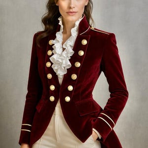 Puede incluir: Chaqueta de terciopelo burdeos con botones y ribetes dorados. La chaqueta tiene un cuello alto y se combina con una blusa blanca con volantes y pantalones color crema. El diseño de la chaqueta recuerda al estilo militar.