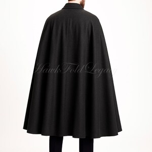 Puede incluir: Una capa de lana gris carb&oacute;n oscuro con cuello estructurado y silueta acampanada de cuerpo entero. La prenda muestra el texto Hawk Fold Legacy en la parte posterior. Se lleva sobre pantalones oscuros y zapatos de vestir de cuero negro.