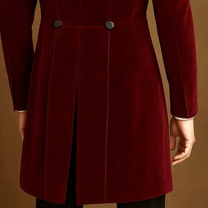 May include: A burgundy velvet coat with black buttons and a pleated back. The coat has long sleeves with white cuffs. The garment is a formal style.